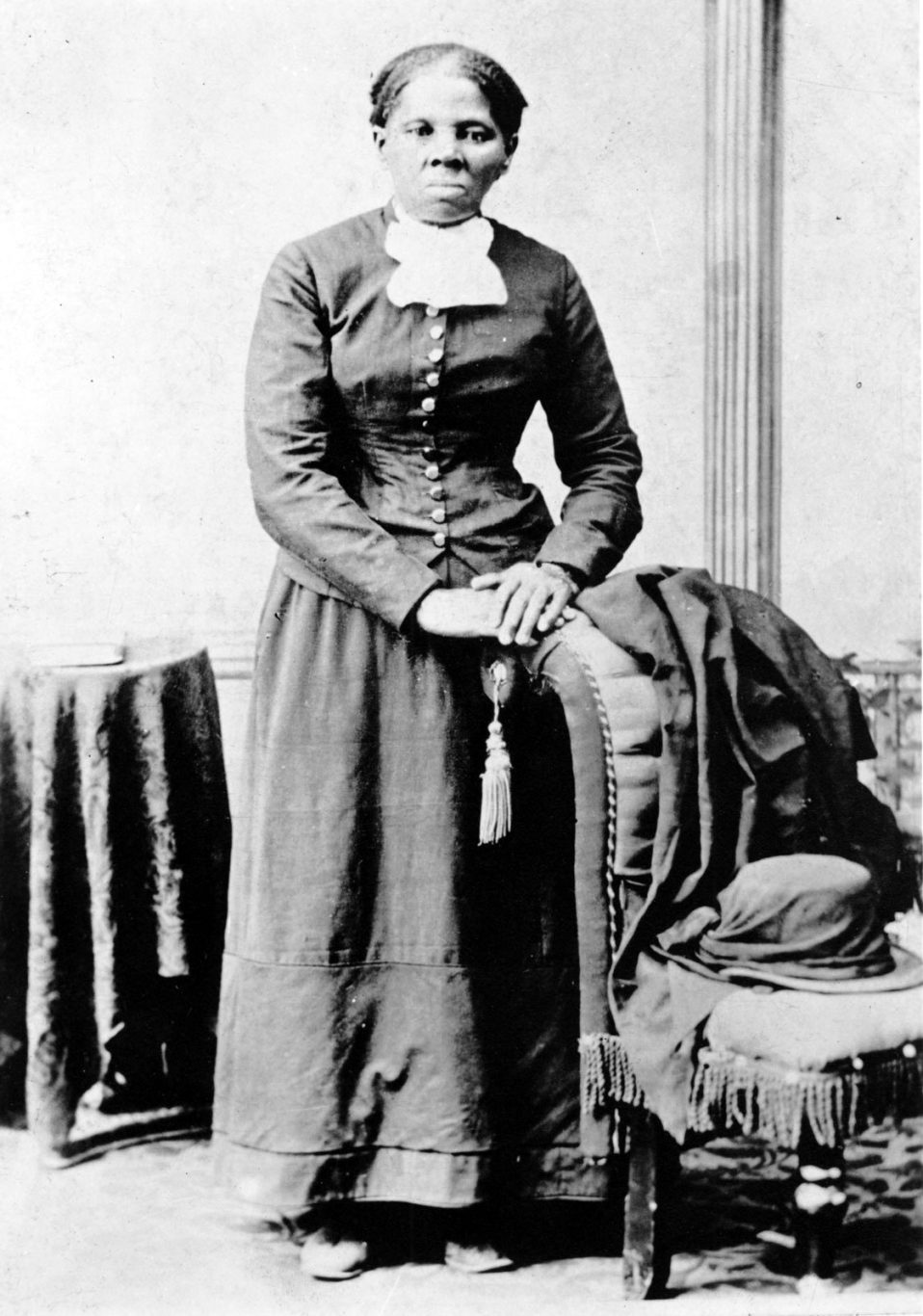 Photograph shows a full-length portrait of Harriet Tubman (1820?-1913) looking directly at the camera with folded hands resting on back of an upholstered chair.