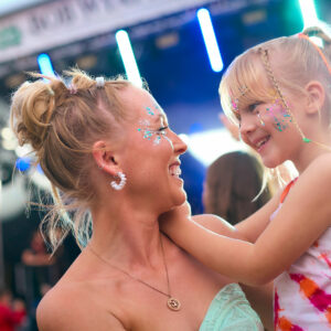 Mother With Daughter Wearing Glitter Having Fun At Outdoor Summer Music Festival