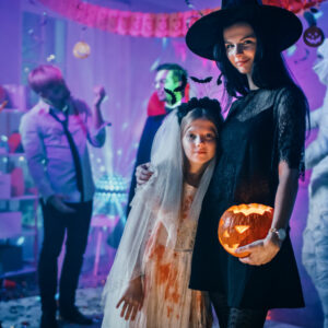 Halloween Costume Party: Portrait of Dazzling Young Witch Holding Burning Pumpkin Head Standing with Smiling Little Girl in a Bloody White Bride Dress. Cool Monster Party with Disco Ball Lights