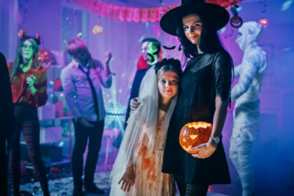 Halloween Costume Party: Portrait of Dazzling Young Witch Holding Burning Pumpkin Head Standing with Smiling Little Girl in a Bloody White Bride Dress. Cool Monster Party with Disco Ball Lights