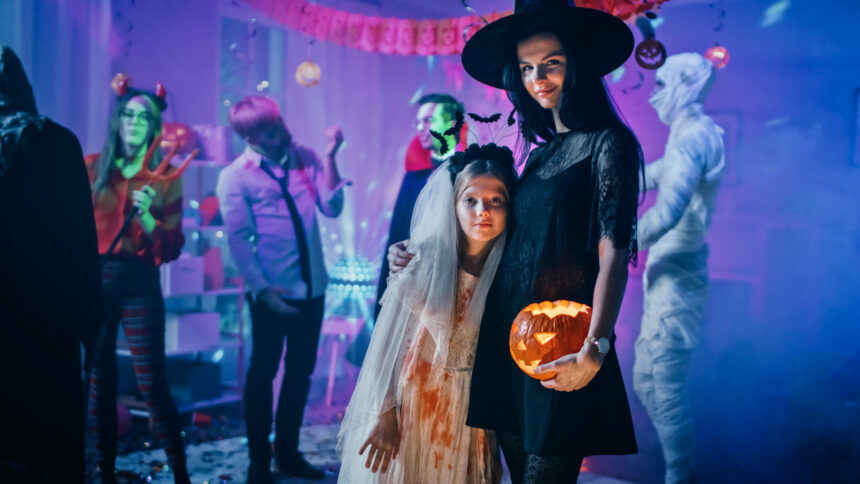Halloween Costume Party: Portrait of Dazzling Young Witch Holding Burning Pumpkin Head Standing with Smiling Little Girl in a Bloody White Bride Dress. Cool Monster Party with Disco Ball Lights