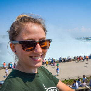 Young happy smiling woman with sunglasses. Beautiful sunny springtime morning at Niagara Falls. People & tourists in background see, hear, and feel the power of the Falls.