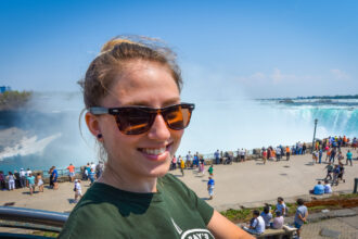 Young happy smiling woman with sunglasses. Beautiful sunny springtime morning at Niagara Falls. People & tourists in background see, hear, and feel the power of the Falls.