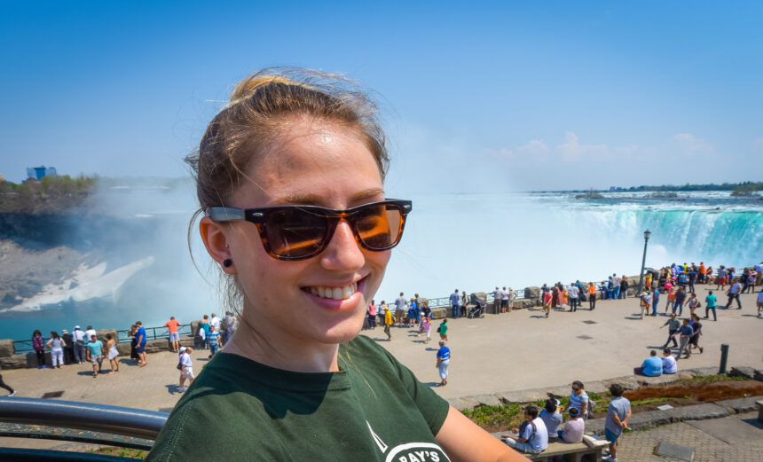 Young happy smiling woman with sunglasses. Beautiful sunny springtime morning at Niagara Falls. People & tourists in background see, hear, and feel the power of the Falls.