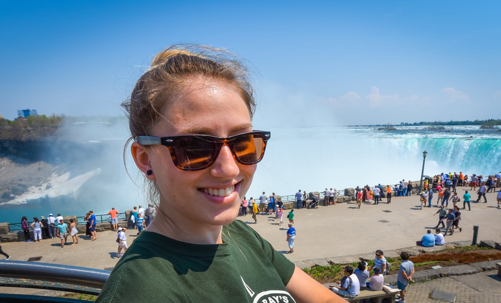 Young happy smiling woman with sunglasses. Beautiful sunny springtime morning at Niagara Falls. People & tourists in background see, hear, and feel the power of the Falls.