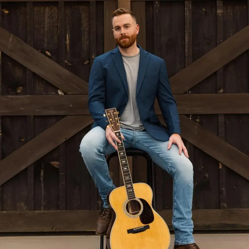 Gavin Ray holding a guitar sitting on a chair with a barndoor in the background