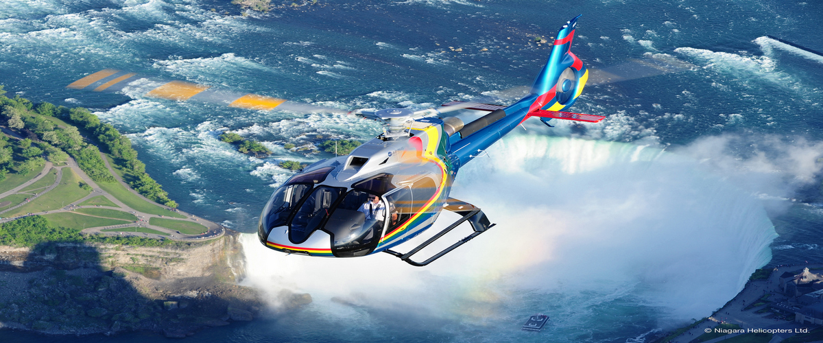 Niagara Falls Helicopter mainly blue in colour with orange, red, and yellow stripes. The backdrop features the Canadian Horsehoe Falls