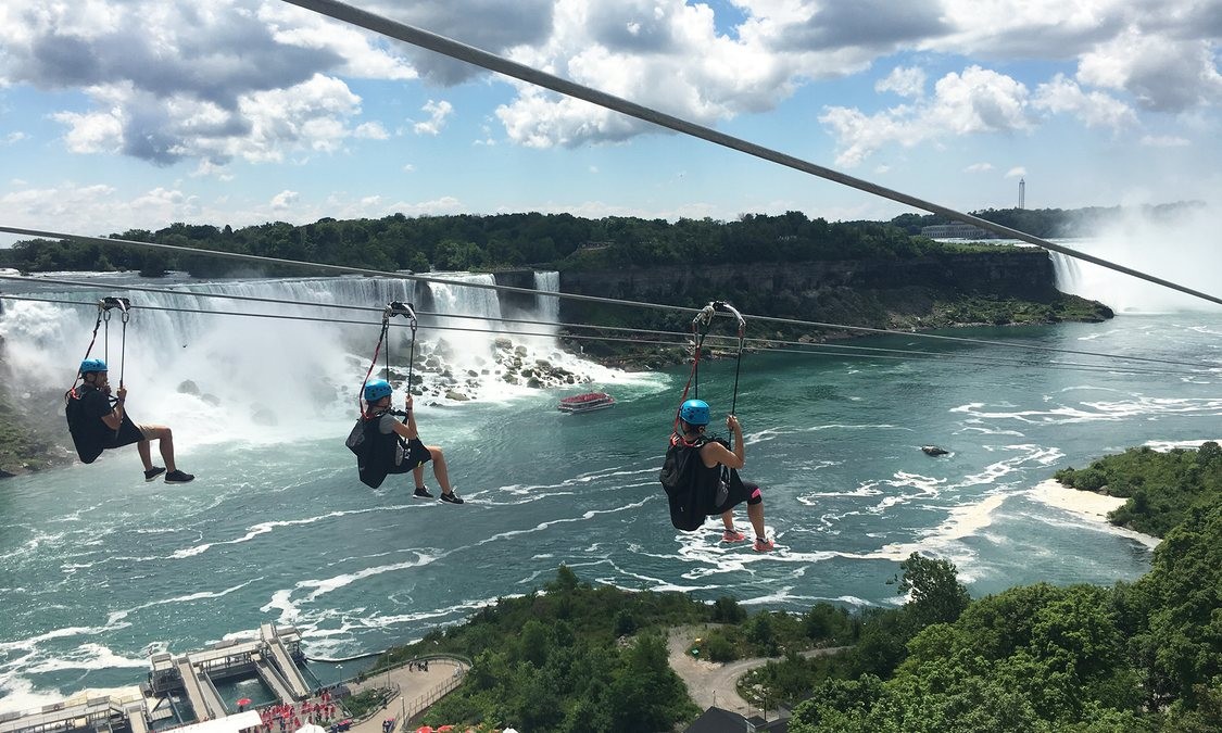 Four passengers riding the Niagara Zipline wearing blue helmets with the American Falls in the background on a clear day. A Maid of the Mist boat is in the bottom right hand corner of the image and foliage surrounding the Gorge and the Niagara Escarpment in the top right hand corner of the image