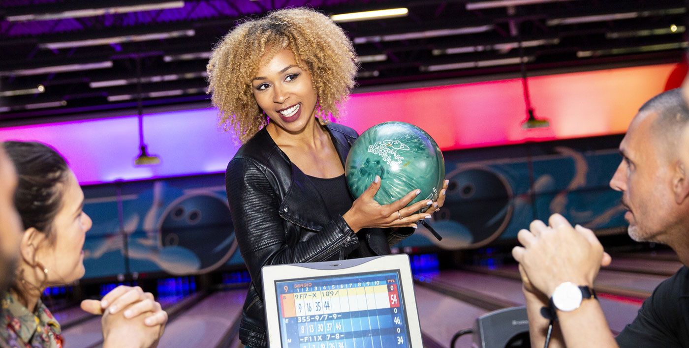 Woman at Strike game zone bowling area motioning to friends with bowling ball in hand