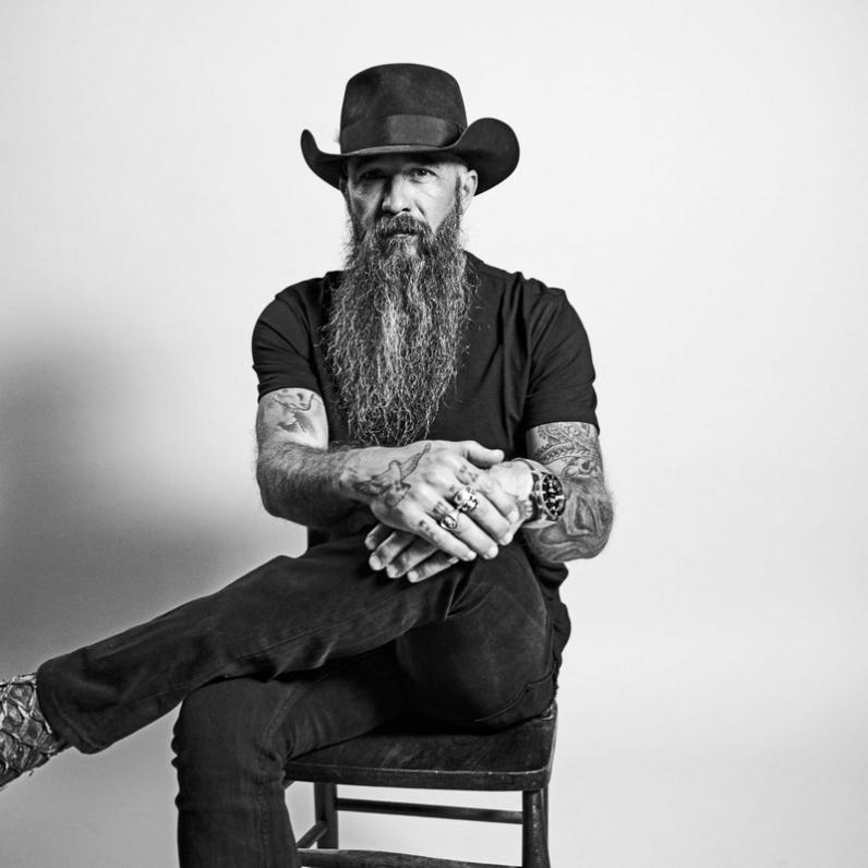 Cody Jinks sitting on a chair with 1 leg crossed over the other, wearing a cowboy hat