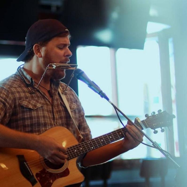 Dave Cassibo wearing a flannel shirt and a backwards baseball cap holding a guitar while holding a harmonica to his lips while standing at a microphone
