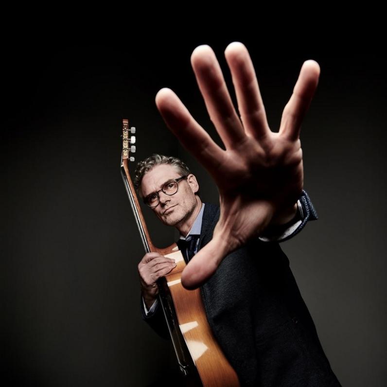 Jesse Cook holding a guitar in his right hand and holding his left hand up