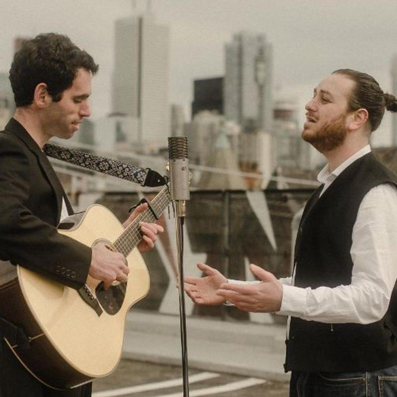 Two men standing, one is holding a guitar, one is singing