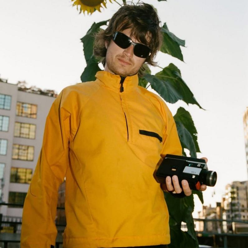 Joe Keery wearing a yellow shirt, sunglasses, and holding a camera