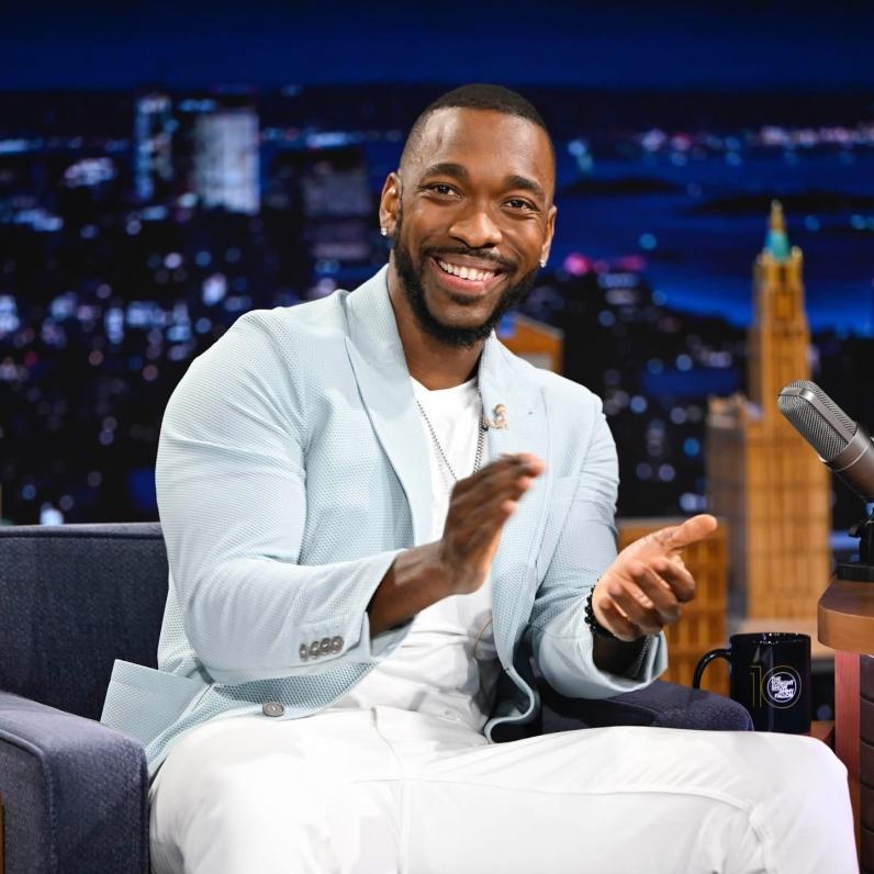 Jay Pharoah sitting on a couch clapping his hands and smiling