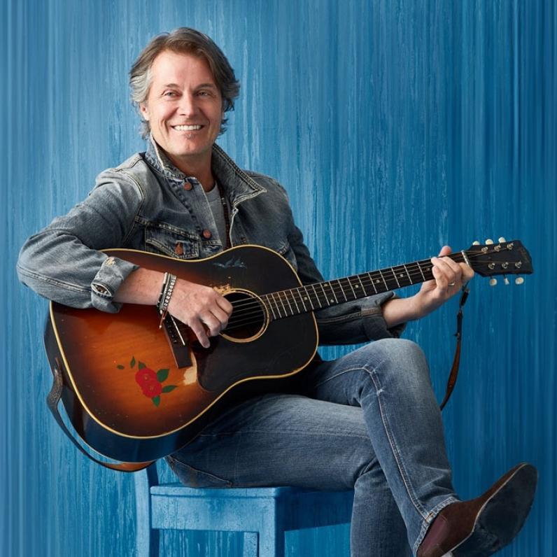 Jim Cuddy sitting on a chair holding a guitar