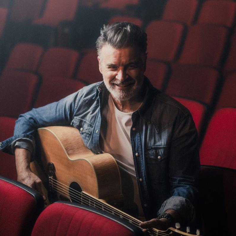 Roch Voisine sitting in audience seats holding a guitar