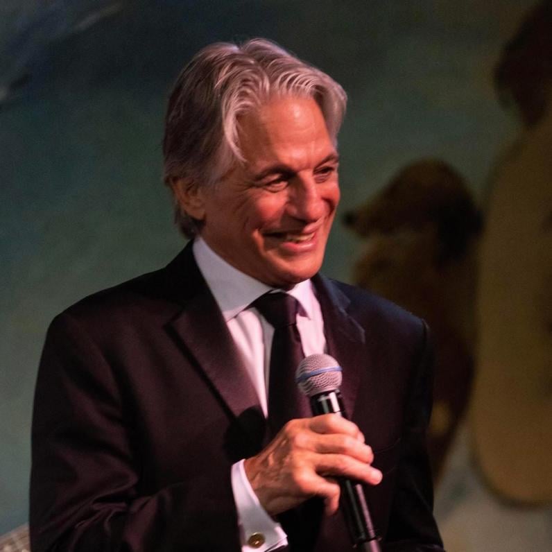 Tony Danza in a suit on stage holding a microphone