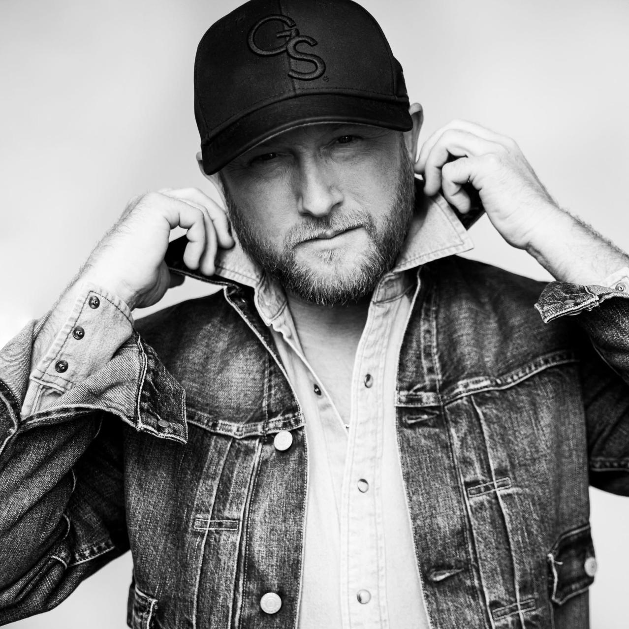 Cole Swindell wearing a jean jacket and a baseball hat, fixing his collar