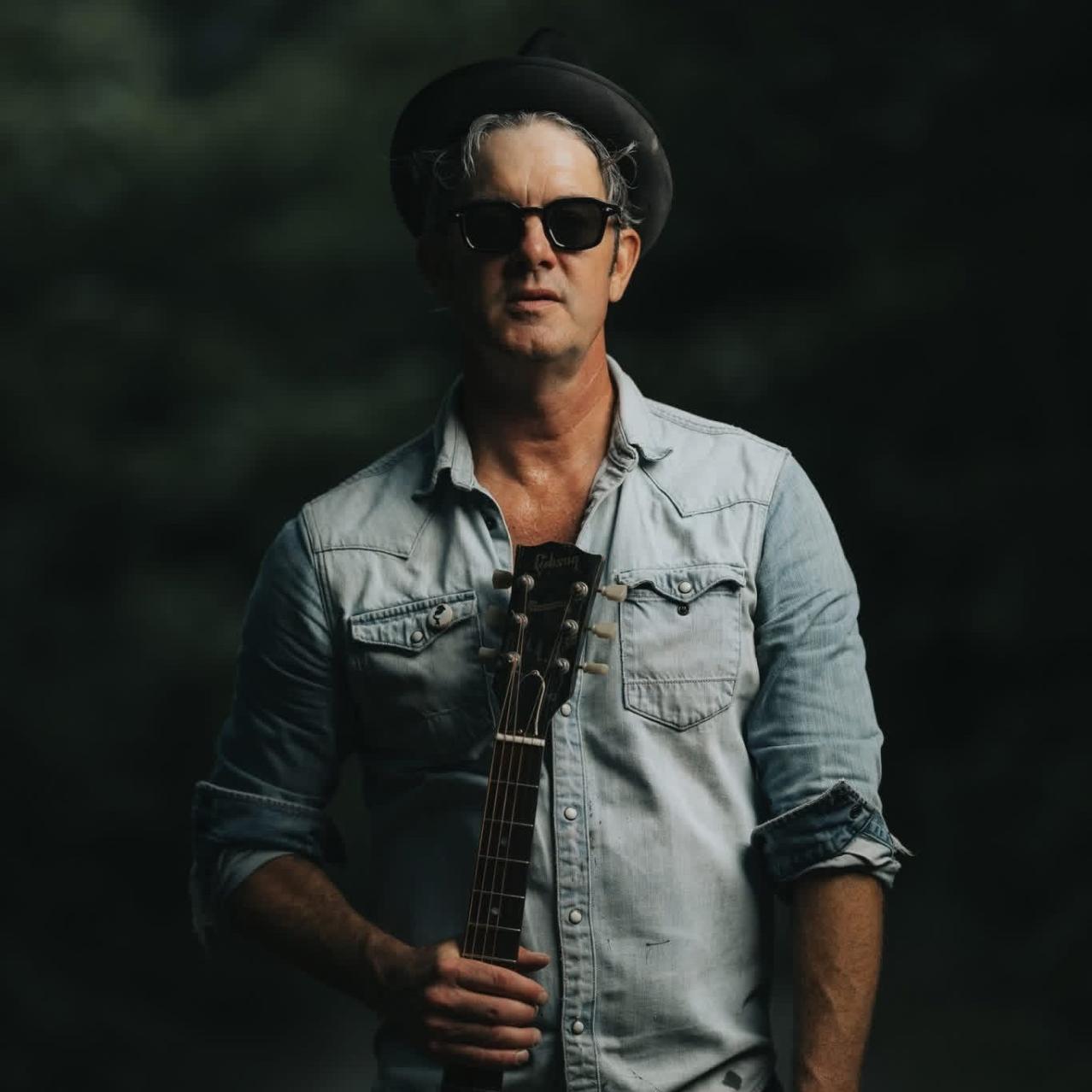 G Love wearing glasses, a hat, and holding a guitar