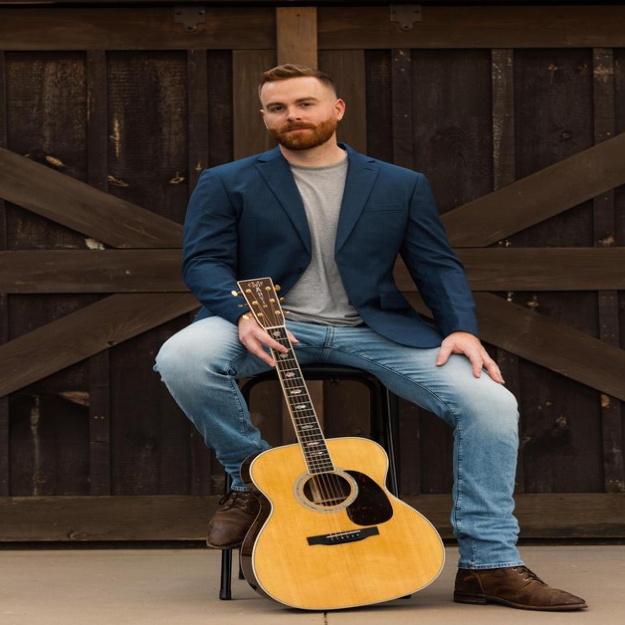 Gavin Ray holding a guitar sitting on a chair with a barndoor in the background