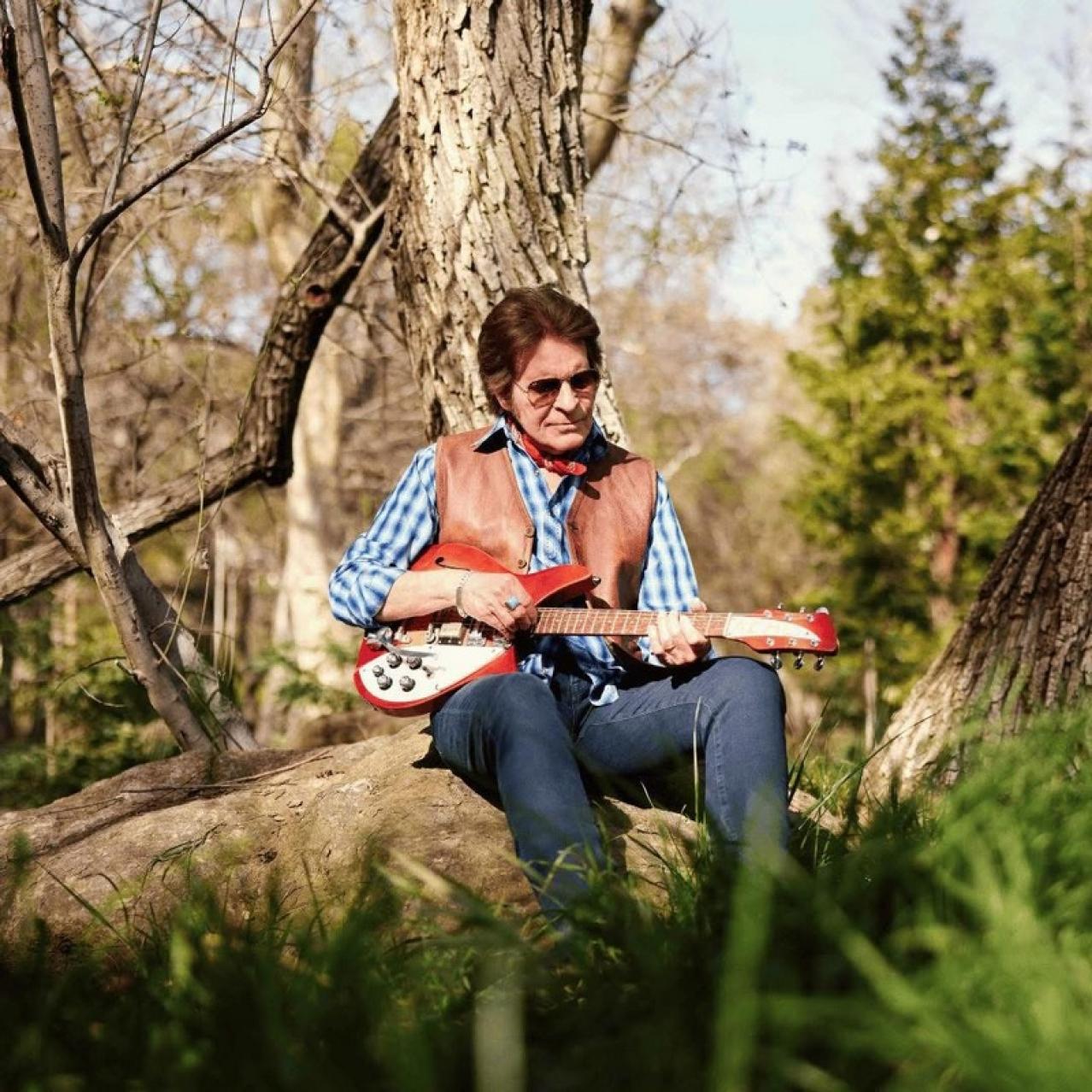John sits on a tree trunk with a guitar in his hands