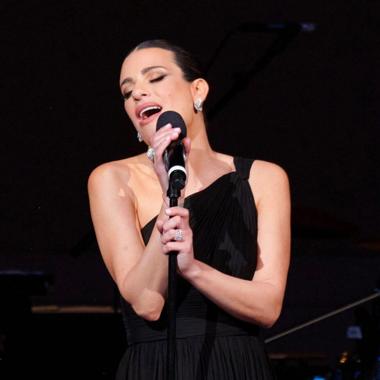Lea Michele is singing with a microphone in her hand in a black one should sleeveless dress