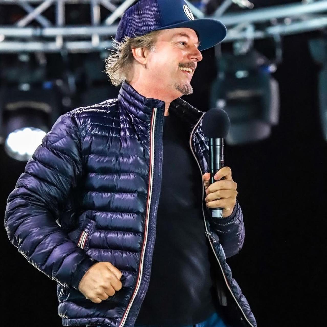 David Spade on stage holding a microphone wearing a baseball hat and fall coat