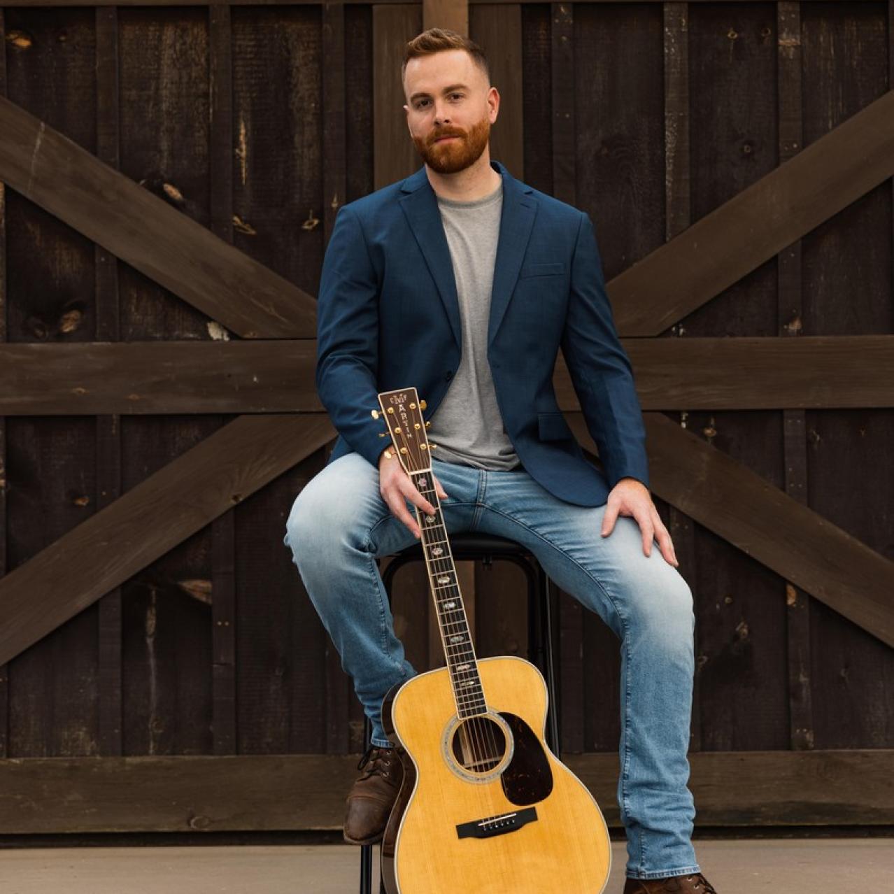 Gavin Ray holding a guitar sitting on a chair with a barndoor in the background
