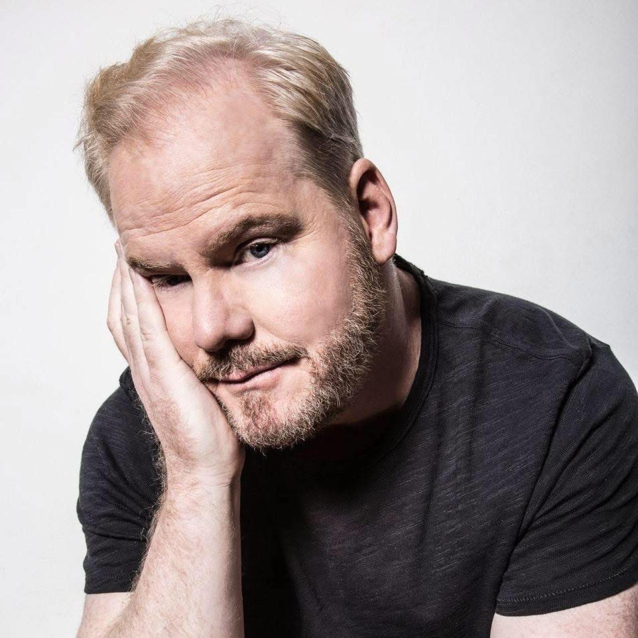 Jim Gaffigan in a black t shirt slouching with his right hand on his cheek
