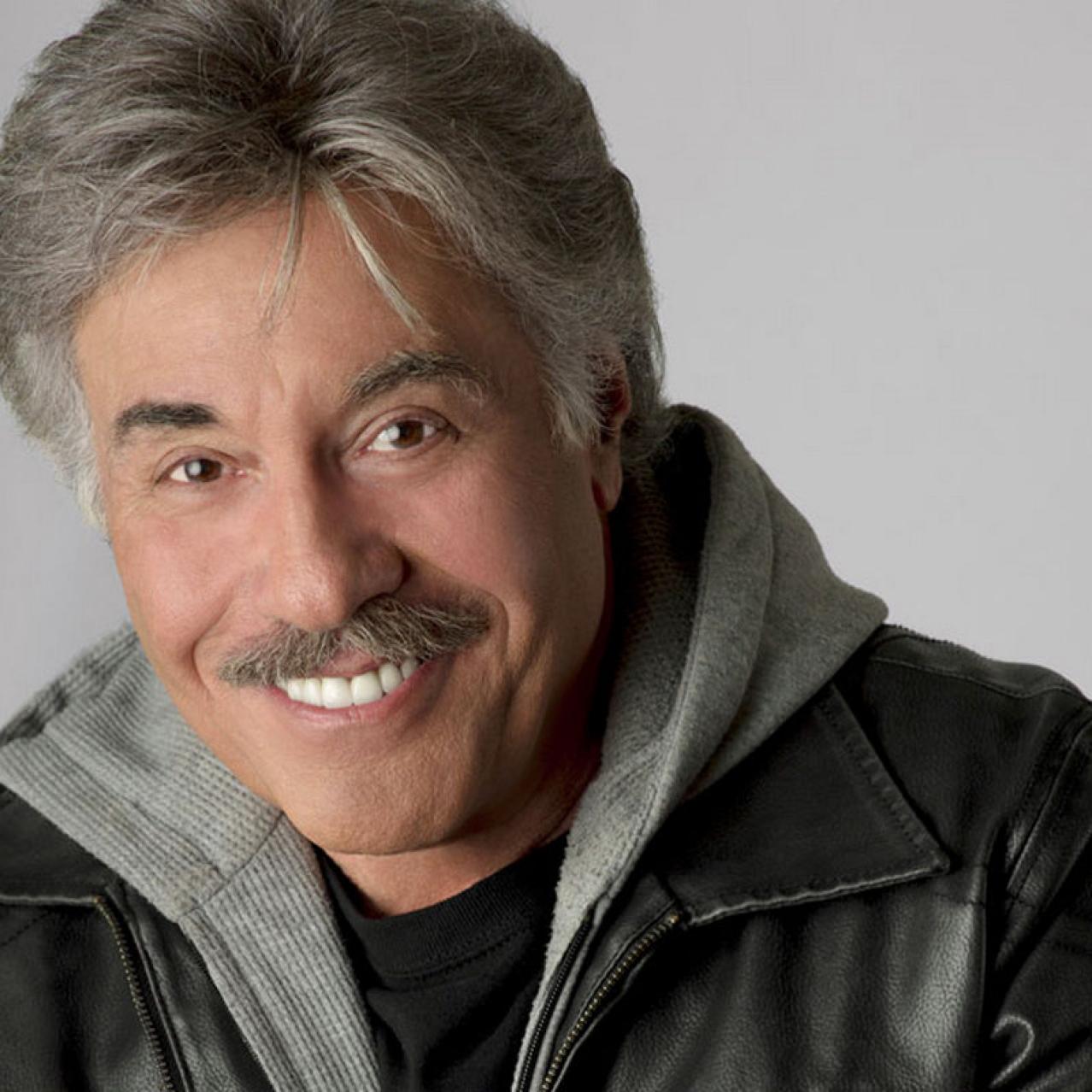Close up headshot of Tony Orlando smiling, wearing a grey hoodie under a black leather jacket.