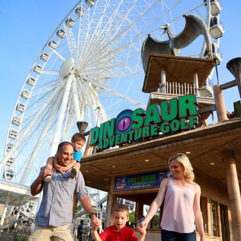 Family with young children walking away from Dino Golf booth with SkyWheel in the background