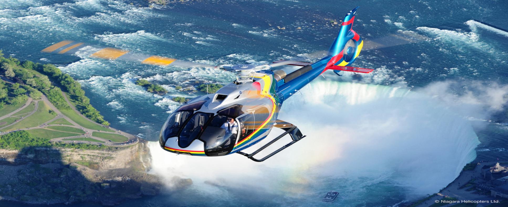 Niagara Falls Helicopter mainly blue in colour with orange, red, and yellow stripes. The backdrop features the Canadian Horsehoe Falls