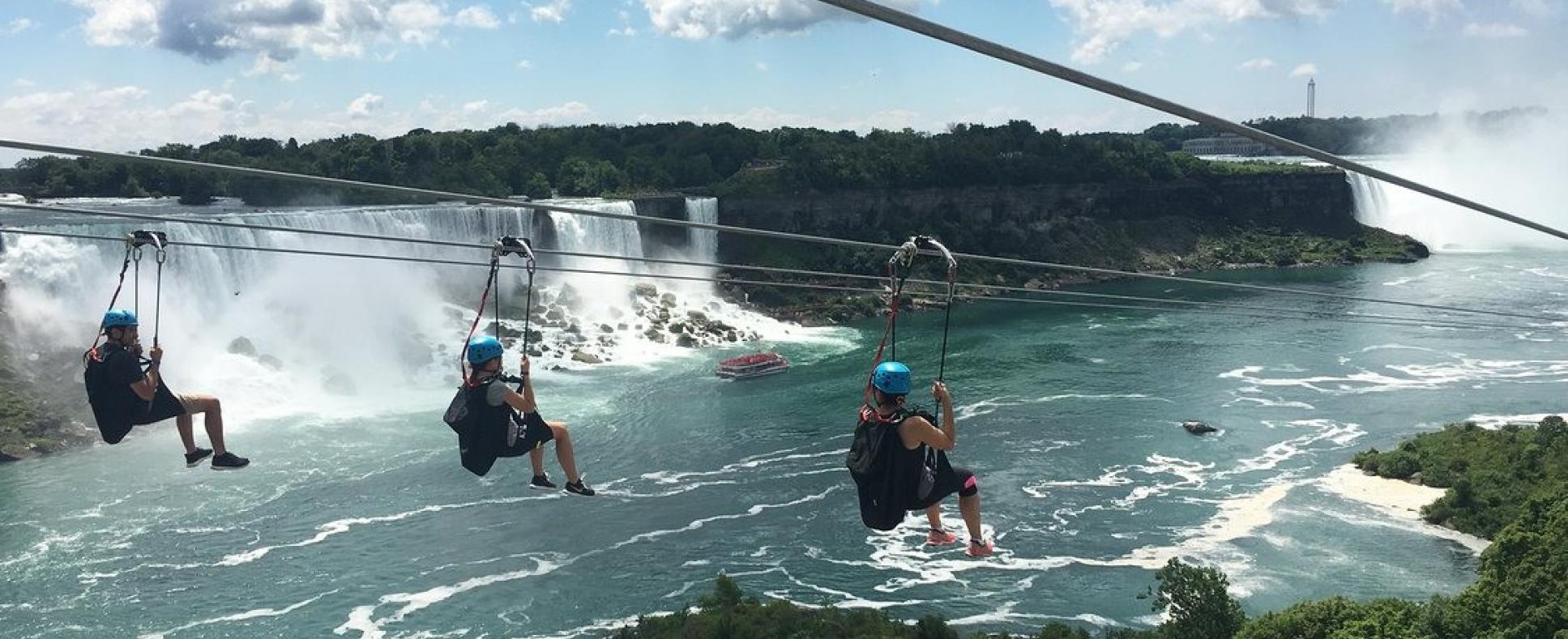 Four passengers riding the Niagara Zipline wearing blue helmets with the American Falls in the background on a clear day. A Maid of the Mist boat is in the bottom right hand corner of the image and foliage surrounding the Gorge and the Niagara Escarpment in the top right hand corner of the image