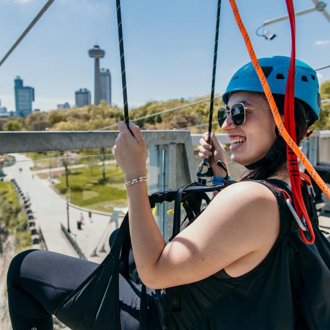 Close up of woman seated in zipline harness wearing a helmet with the Skylon and Fallsview in the horizon